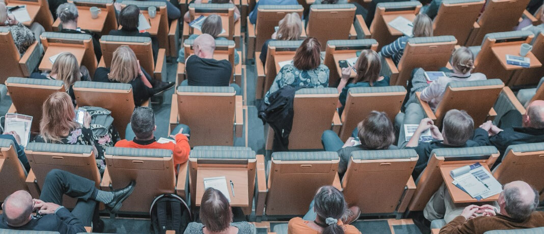 A lecture hall filled with people seated in wooden chairs, viewed from above. Many attendees hold papers, and some have drinks on small desks attached to the chairs. The atmosphere suggests a formal presentation or academic setting.