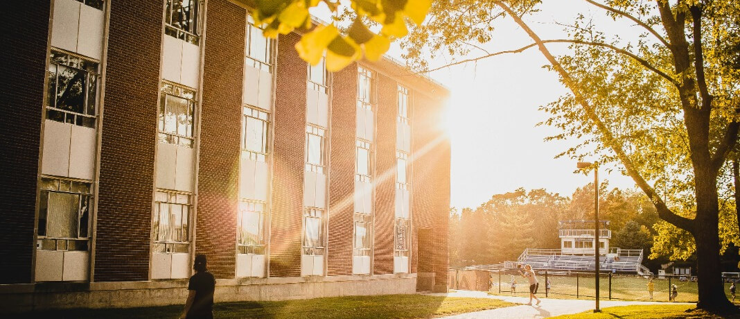 A large brick building with rows of windows is bathed in golden sunset light. Two people walk along a path near the building. In the background, a sports field and bleachers are visible, surrounded by trees.