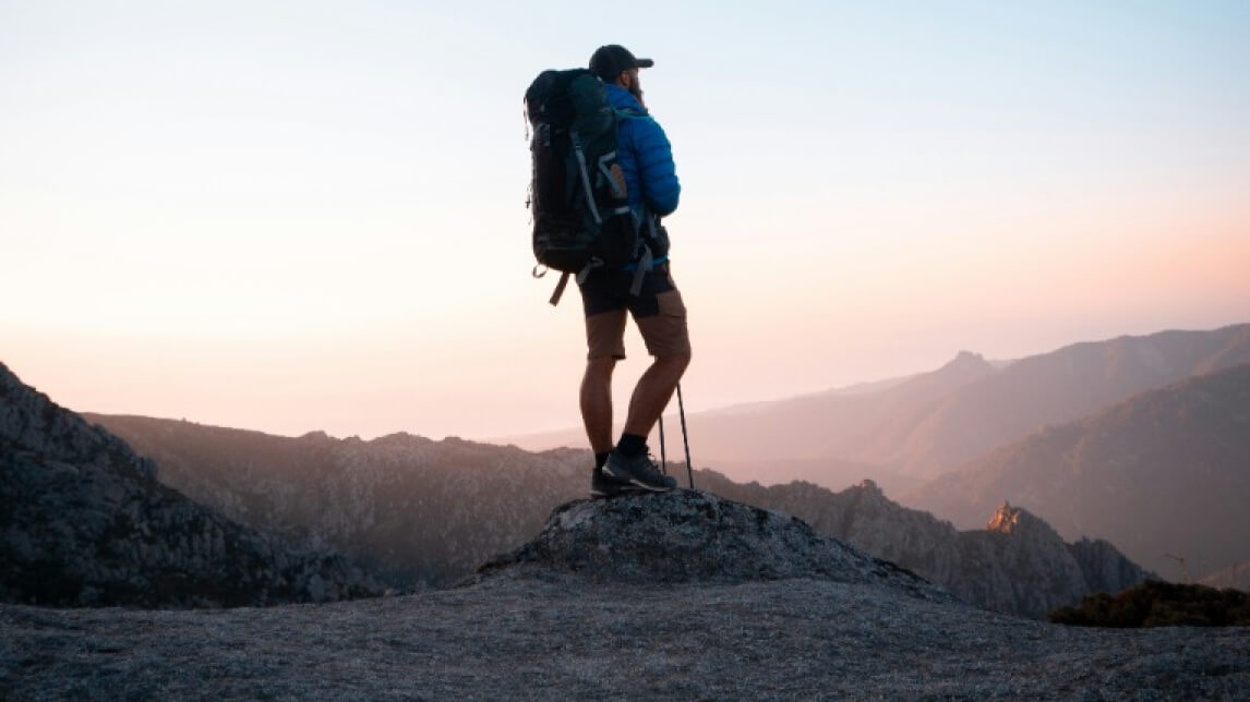A person in outdoor gear, including a blue jacket and a cap, stands on a rocky peak with a large backpack and hiking poles. The backdrop features a vast mountain landscape under a clear, pastel-colored sky.