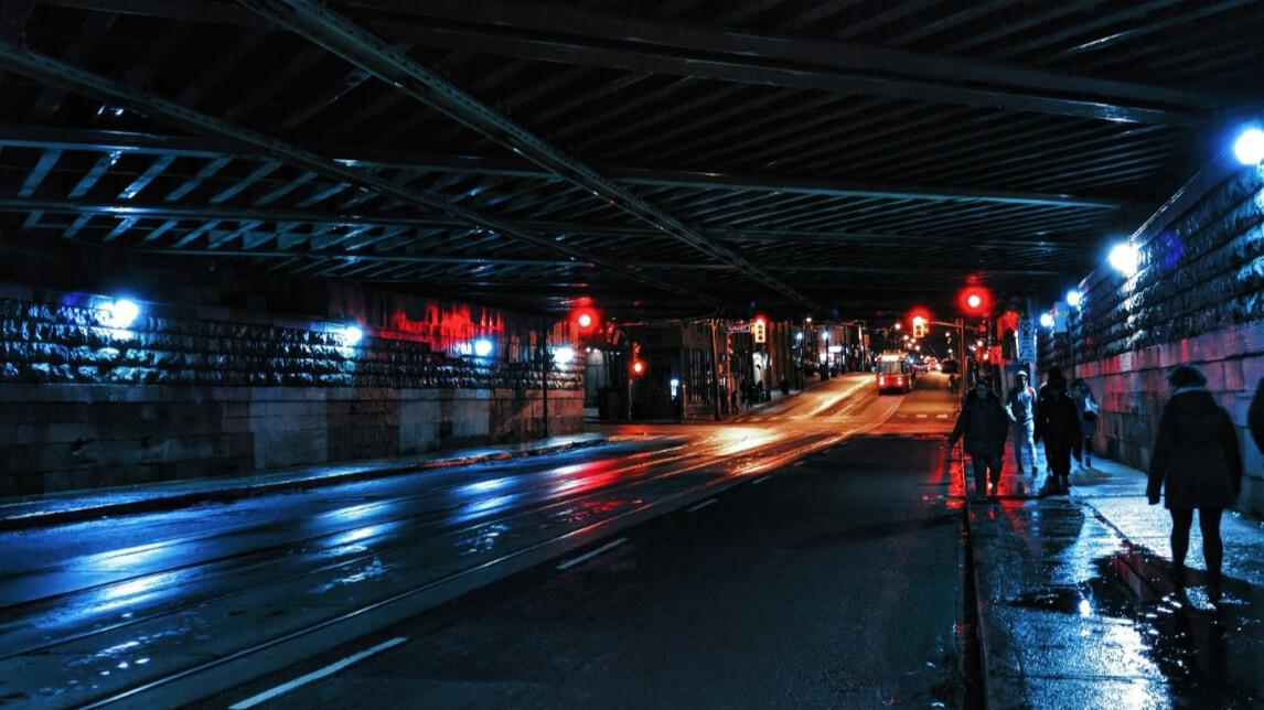 A dimly lit urban underpass at night with wet roads reflecting streetlights. Cars and pedestrians are visible in the distance, illuminated by red and white lights. The structures overhead beams and stone walls create a tunnel-like atmosphere.