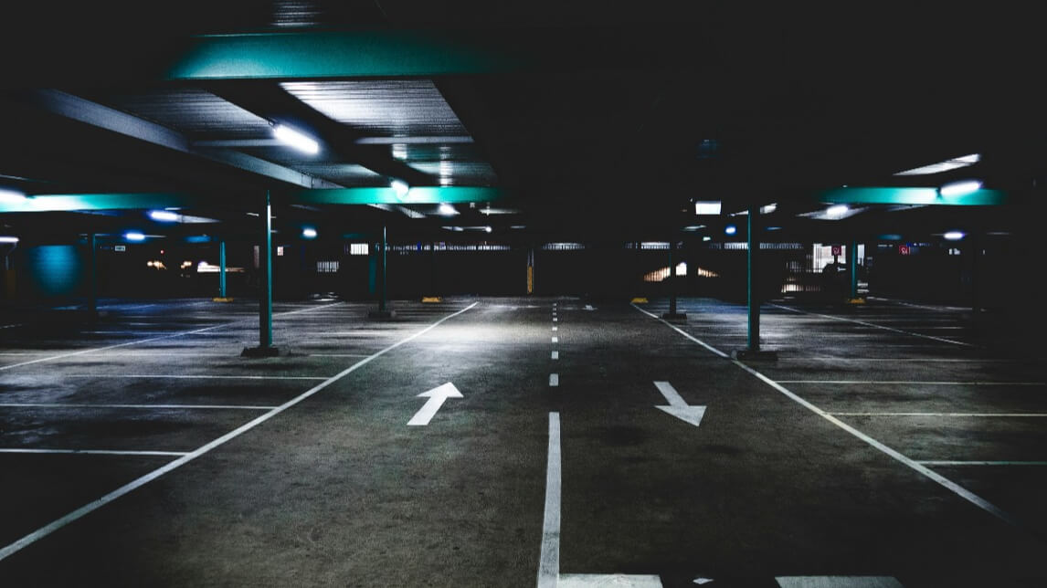 A dimly lit, empty underground parking lot with painted arrows on the concrete floor guiding traffic. The black ceiling has evenly spaced lights, casting a soft glow on the parking spaces and creating an urban, industrial atmosphere.