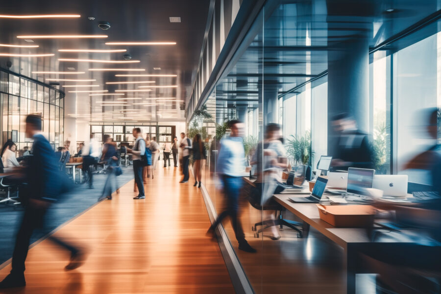 A modern office with a long corridor and glass walls. People are walking and working at desks with computers. The atmosphere is busy and dynamic, with bright lighting and reflections on the floor.