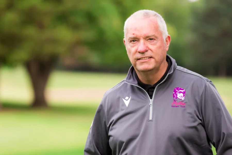 A man with short gray hair stands in a park, wearing a gray sports jacket with a logo on the left side. The background features lush greenery and blurred trees, suggesting a natural, outdoor setting.
